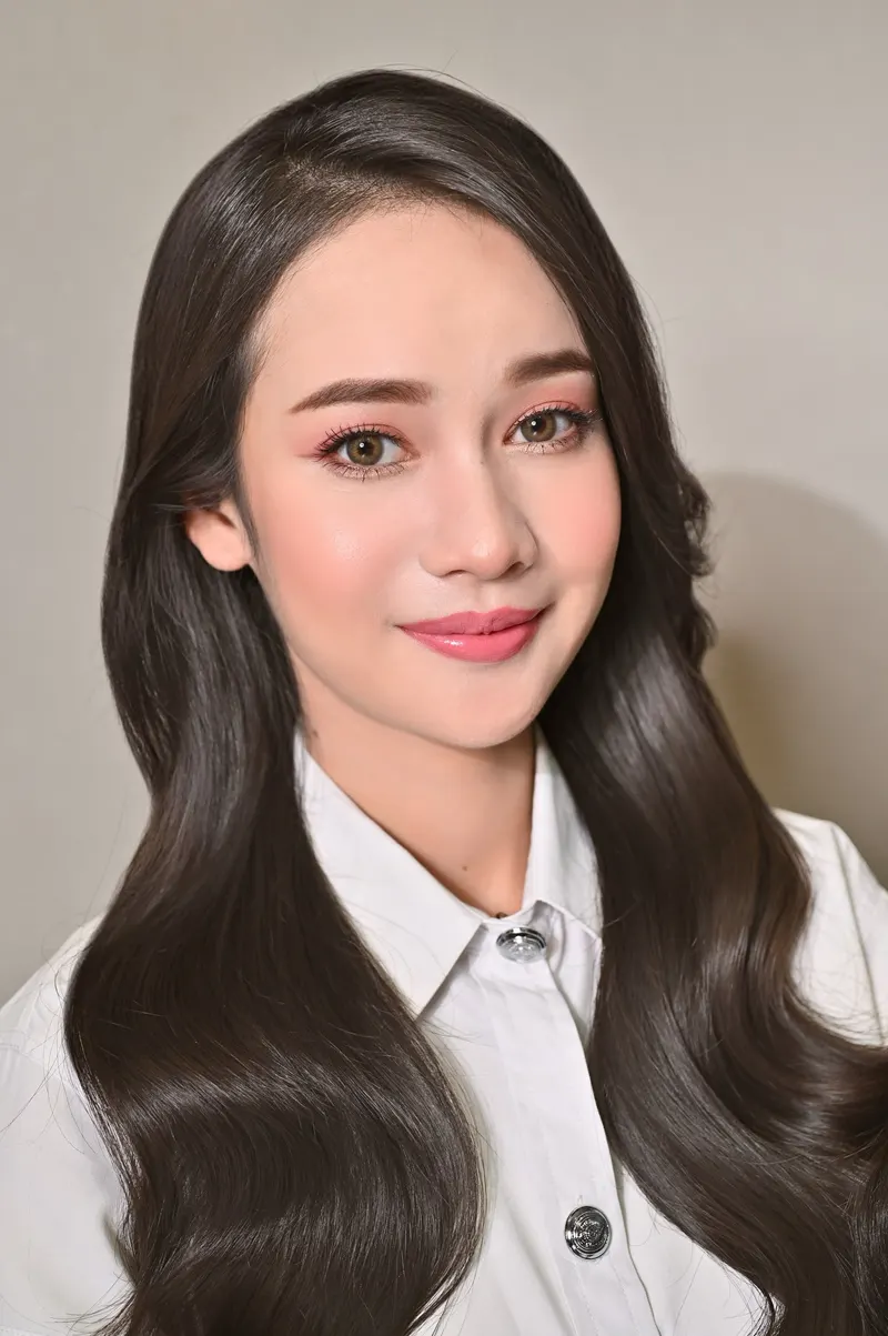 ผมดำยาวลอนใหญ่ปล่อยทั้งสองด้าน แต่งตาชมพูพีช ปัดแก้มพีช ลิปสีกุหลาบ ในเสื้อนักศึกษาสีขาว พื้นหลังสีเทาเรียบ