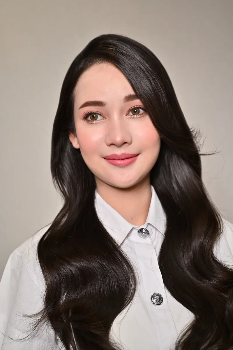 ผมดำยาวลอนใหญ่เกลี่ยแสงเงา แต่งหน้าโทนชมพูนู้ด ลิปสีชมพูกุหลาบ ในเสื้อนักศึกษาสีขาว ยิ้มบาง ฉากหลังสีเทา