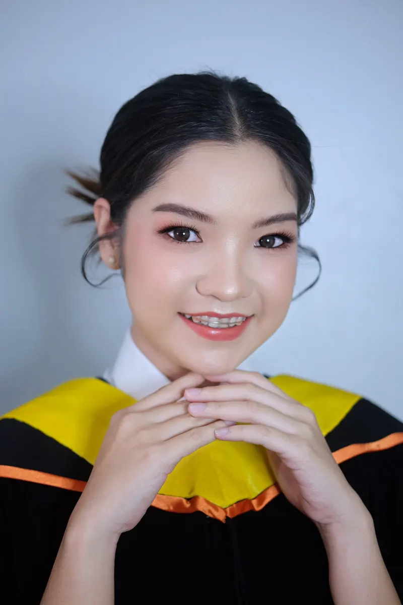 บัณฑิตสวมครุยดำผ้าคาดสีเหลืองขอบส้ม ประสานมือใต้คาง ผมรวบต่ำปล่อยปอยข้างหน้า แต่งหน้าสดใสโทนพีชชมพู ฉากสีฟ้าเทา