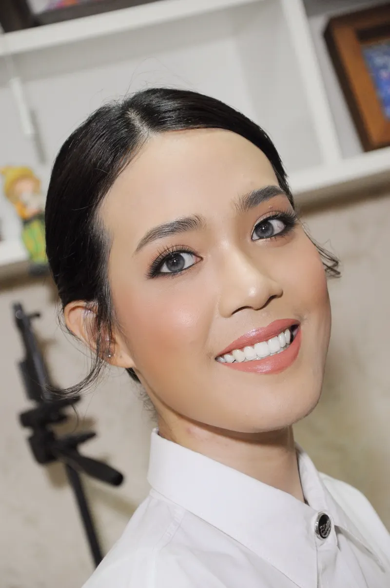 ใบหน้าเอียงยิ้มเห็นฟัน ผมรวบตึงเรียบ ขนตาและอายไลเนอร์เน้นตาโต ลิปสีนู้ดส้ม ในเสื้อนักศึกษาสีขาว ฉากชั้นวางของ