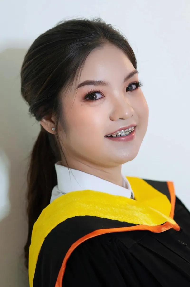บัณฑิตเงยหน้ายิ้มเห็นเครื่องมือจัดฟัน ผมรวบหางม้าต่ำ ขนตาหนา ปัดแก้มพีชชมพู ครุยดำผ้าคาดสีเหลืองขอบส้ม ฉากสีขาว