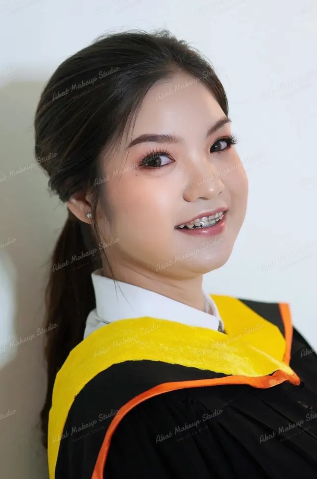 บัณฑิตเงยหน้ายิ้มเห็นเครื่องมือจัดฟัน ผมรวบหางม้าต่ำ ขนตาหนา ปัดแก้มพีชชมพู ครุยดำผ้าคาดสีเหลืองขอบส้ม ฉากสีขาว