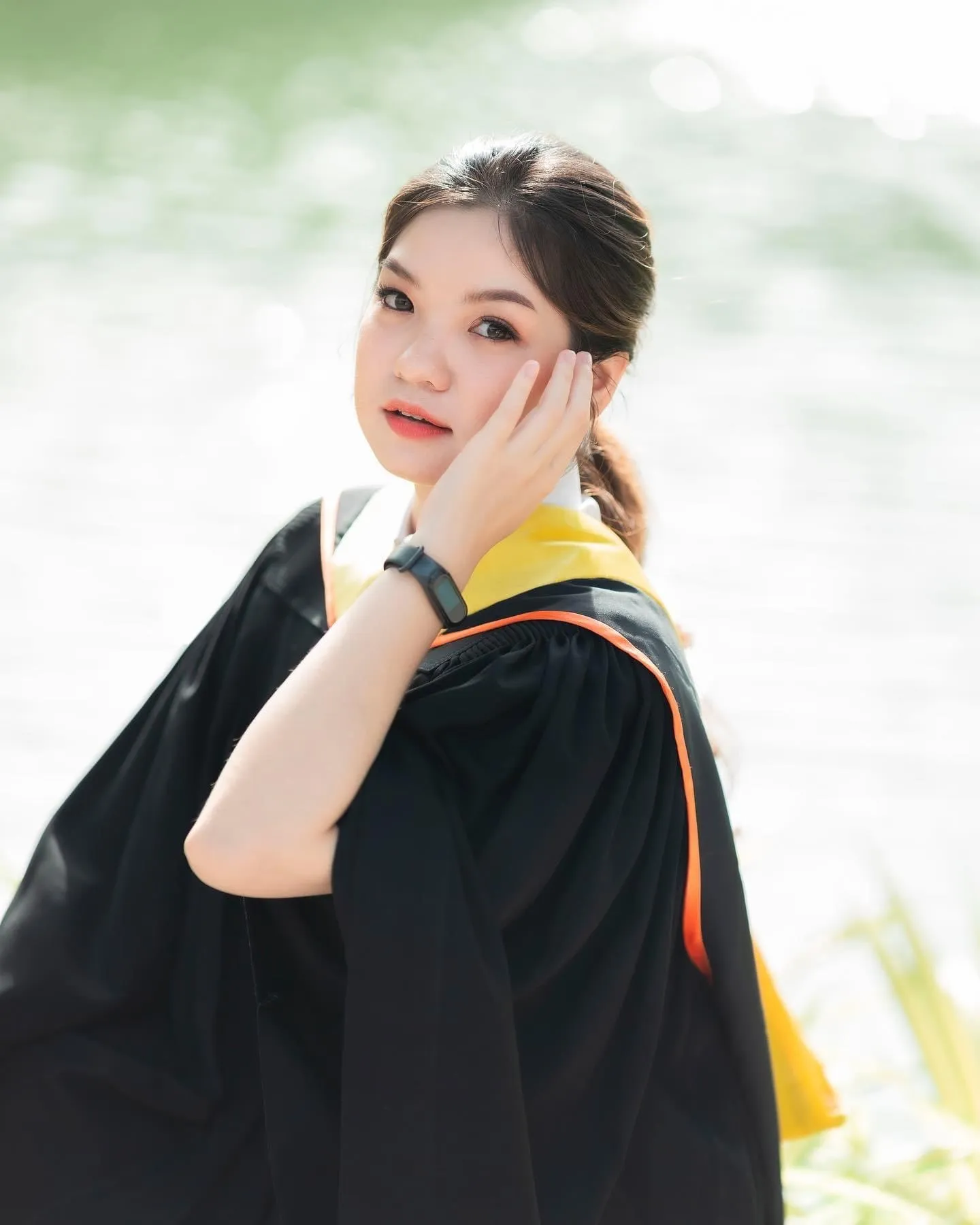 บัณฑิตหญิงริมน้ำในแสงแดดสว่าง ผมรวบต่ำ เอามือแตะแก้ม สวมครุยดำผ้าคาดสีเหลืองขอบส้มกับสมาร์ตวอทช์สีดำ แต่งหน้าโทนพีชธรรมชาติ