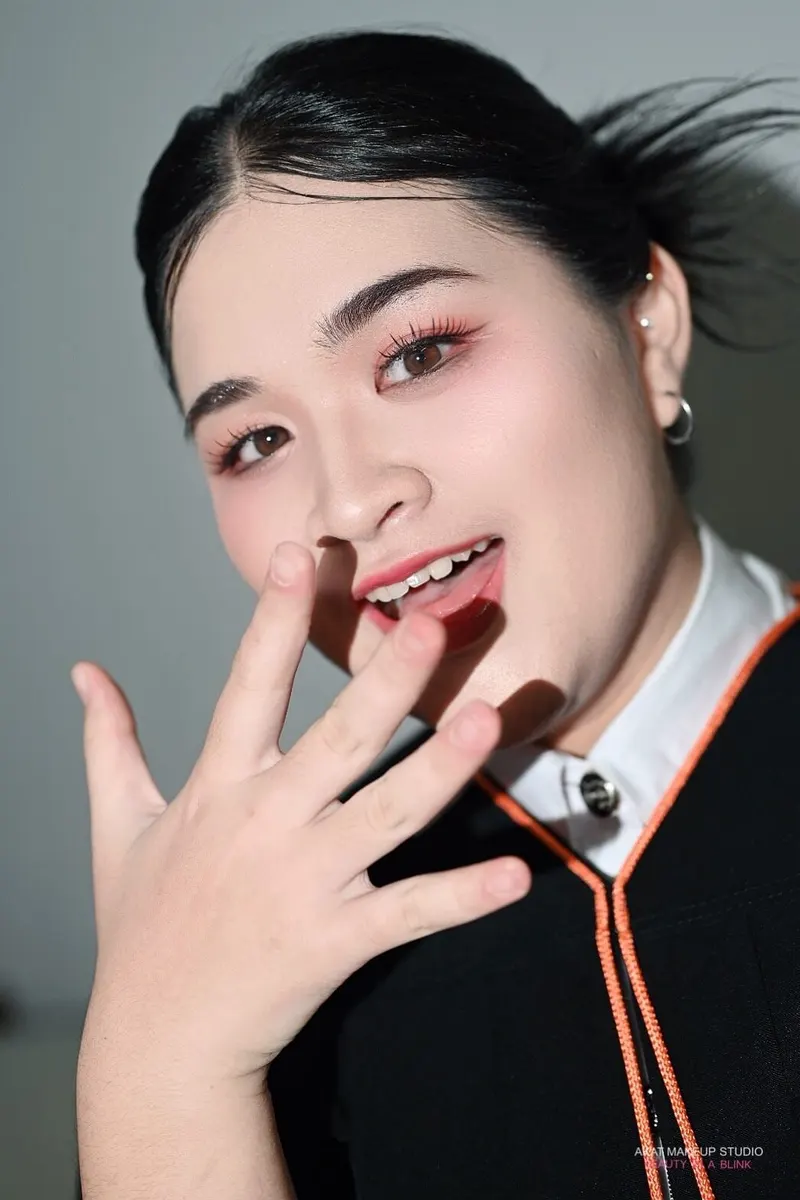 บัณฑิตสวมครุยดำขอบส้ม ยกมือแตะริมฝีปาก ผมสีดำรวบหางสูงปล่อยปอยปลิว แต่งตาชมพูแดงและลิปสีชมพูส้ม ลายน้ำสตูดิโอมุมล่าง