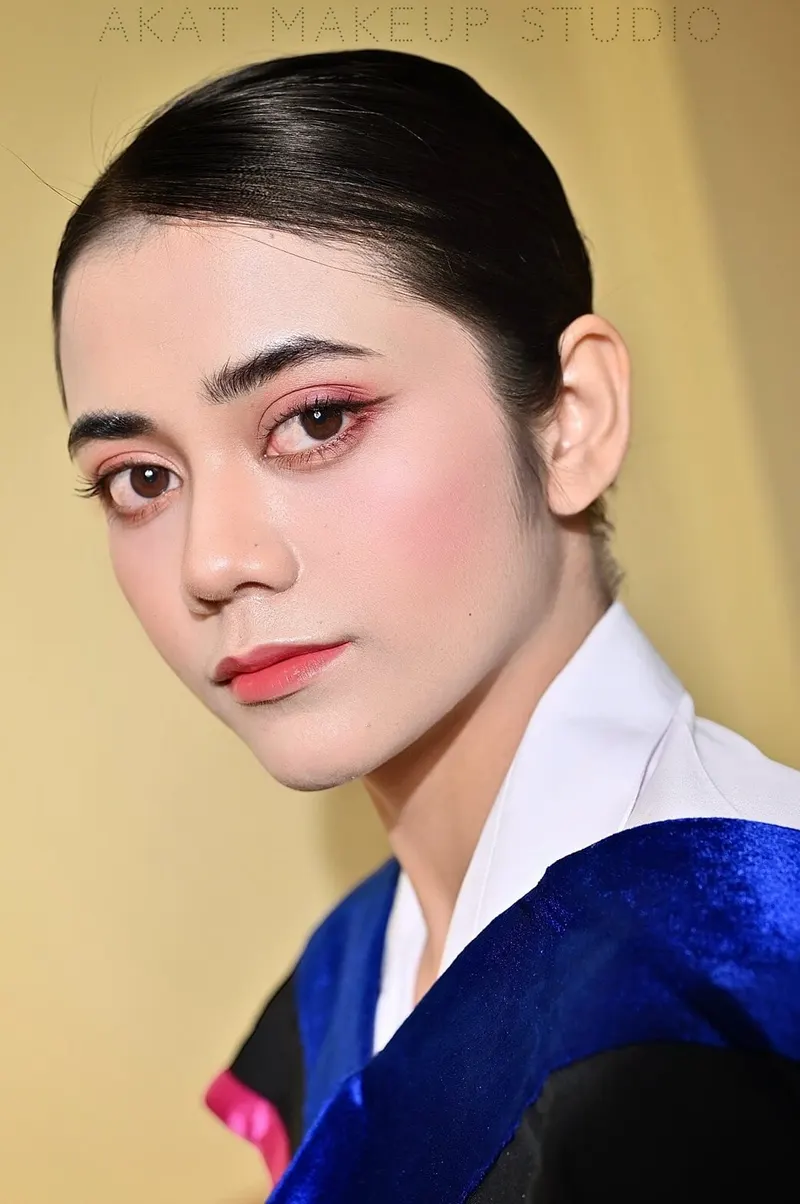 ภาพใกล้ใบหน้าด้านข้าง ผมรวบตึงดำเรียบ คิ้วเข้ม แต่งตาโทนแดงชมพู ปัดแก้มชมพู ลิปสีส้มคอรัล ครุยดำผ้าคาดสีน้ำเงินกำมะหยี่