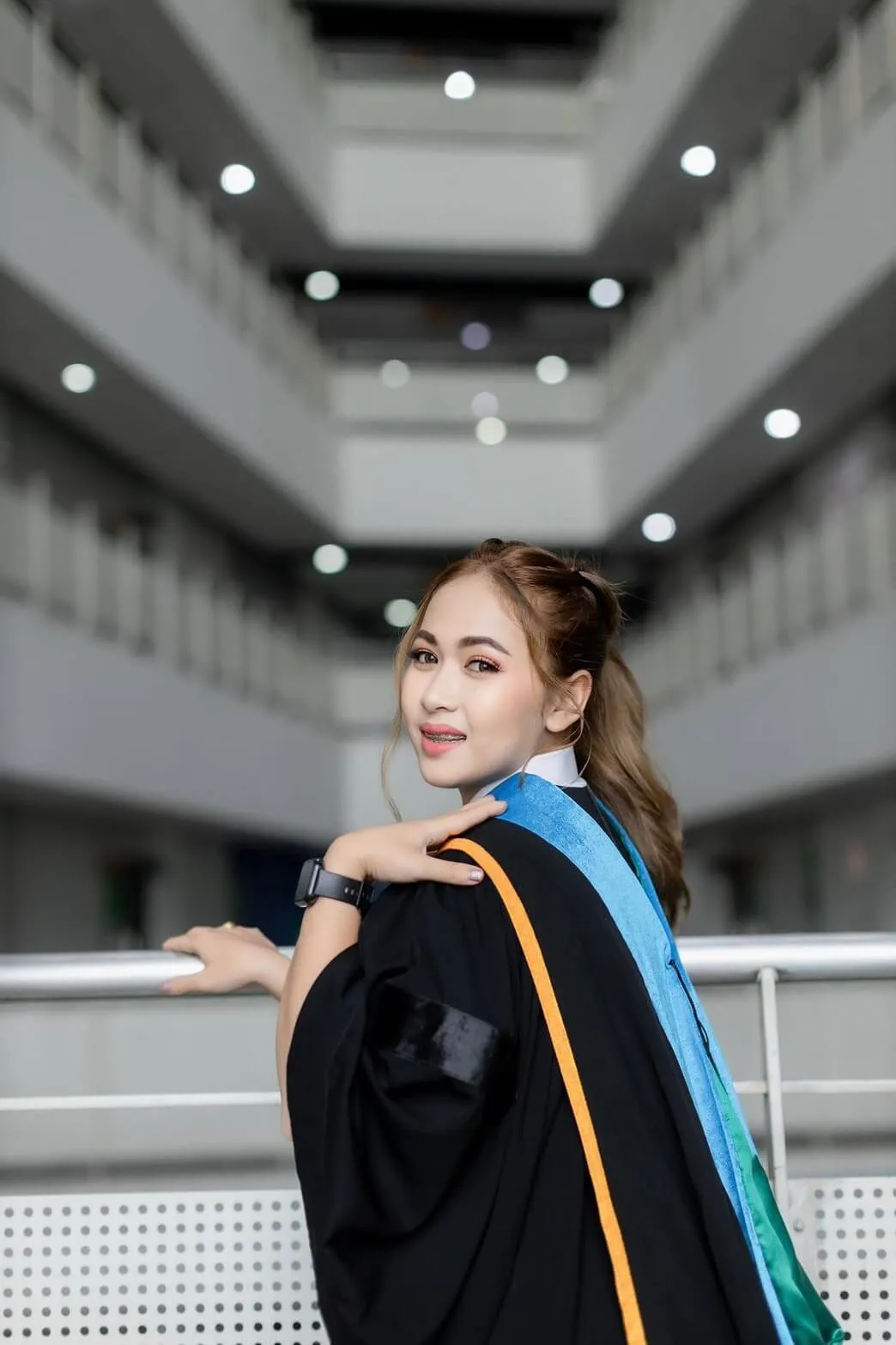 บัณฑิตสาวหันข้างมองกล้องบนระเบียงอาคารโถงสูง ผมรวบหางม้าสูง สวมครุยดำผ้าคาดสีฟ้าเขียวขอบส้ม แต่งหน้าโทนส้มพีช นาฬิกาสีดำที่ข้อมือ