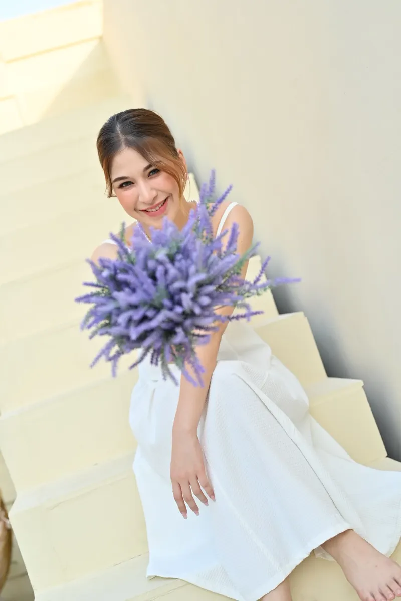 นางแบบนั่งบนบันไดสีครีม สวมเดรสสีขาวสายเดี่ยว ถือช่อลาเวนเดอร์สีม่วง ยิ้มสดใส ผมรวบเก็บ แต่งหน้าสว่างโทนชมพูพีช