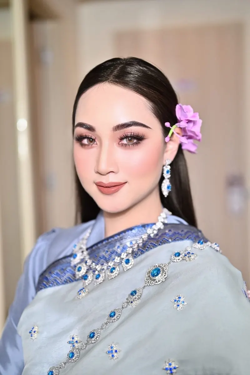ลุคชุดไทยสไบสีฟ้าอ่อนประดับพลอยน้ำเงิน ผมดำรวบประดับกล้วยไม้สีม่วงข้างหู แต่งตาโทนชมพูน้ำตาล ขนตายาว ลิปสีนู้ดส้ม Akat Makeup Studio