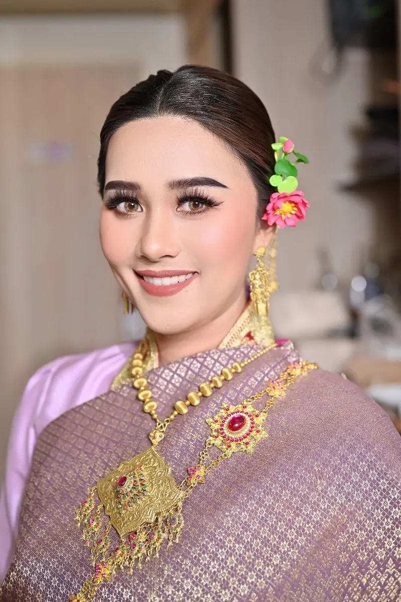 ลุคชุดไทยสไบสีม่วงลายทอง ผมรวบประดับดอกไม้สีชมพูข้างหู เครื่องประดับทองประดับทับทิม แต่งตาโทนน้ำตาลเข้ม ขนตาหนา ยิ้มกว้าง