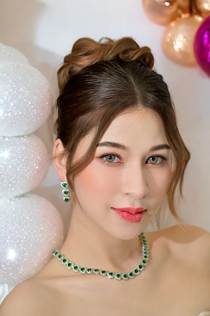 ภาพถ่ายใกล้ธีมงานฉลอง ผมเกล้ามวยสูงปล่อยปอยข้างหน้า สวมสร้อยคอและต่างหูมรกต แต่งตาโทนพีช ลิปชมพูส้ม ฉากลูกโป่งสีขาวและทอง