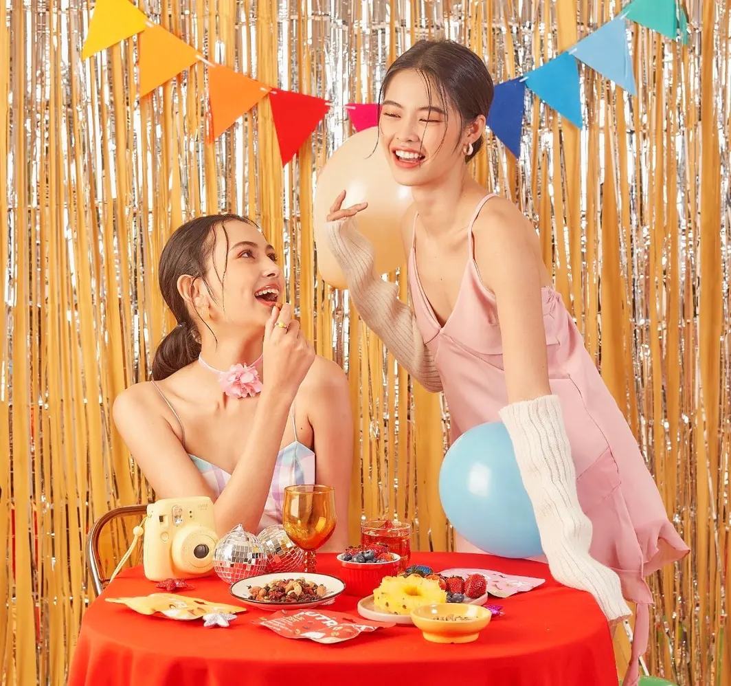 นางแบบสองคนหัวเราะที่โต๊ะผ้าสีแดง ฉากฟอยล์เงินทองและธงราว ถือลูกโป่งและกล้องฟิล์ม แต่งหน้าสดใสโทนชมพูส้ม สไตล์แคมเปญปาร์ตี้