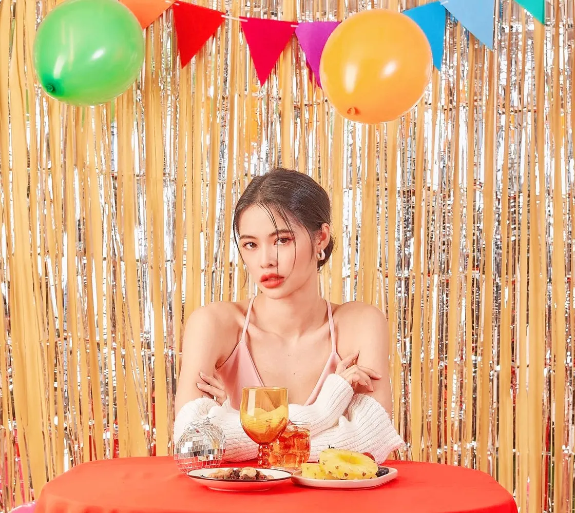 นางแบบผมเกล้าสูงนั่งกอดอกที่โต๊ะผ้าสีแดง มีผลไม้และเครื่องดื่ม ฉากฟอยล์ทองกับลูกโป่งและธงราว แต่งตาโทนส้มชมพู ลิปแดง
