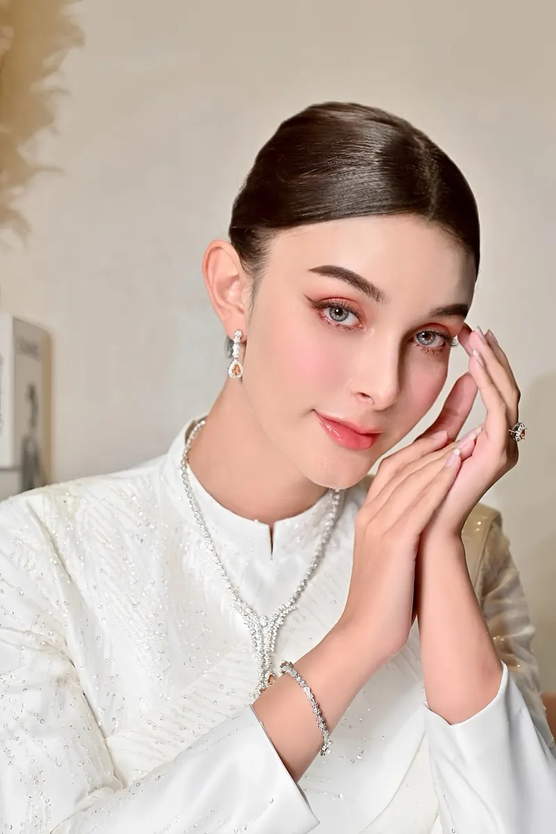 เจ้าสาวในชุดไทยสีขาวปักเลื่อม สวมสร้อยคอเพชร ประสานมือแนบแก้ม แต่งตาโทนส้มพีชและปัดแก้มชมพูอมส้ม ผมรวบเรียบตึง