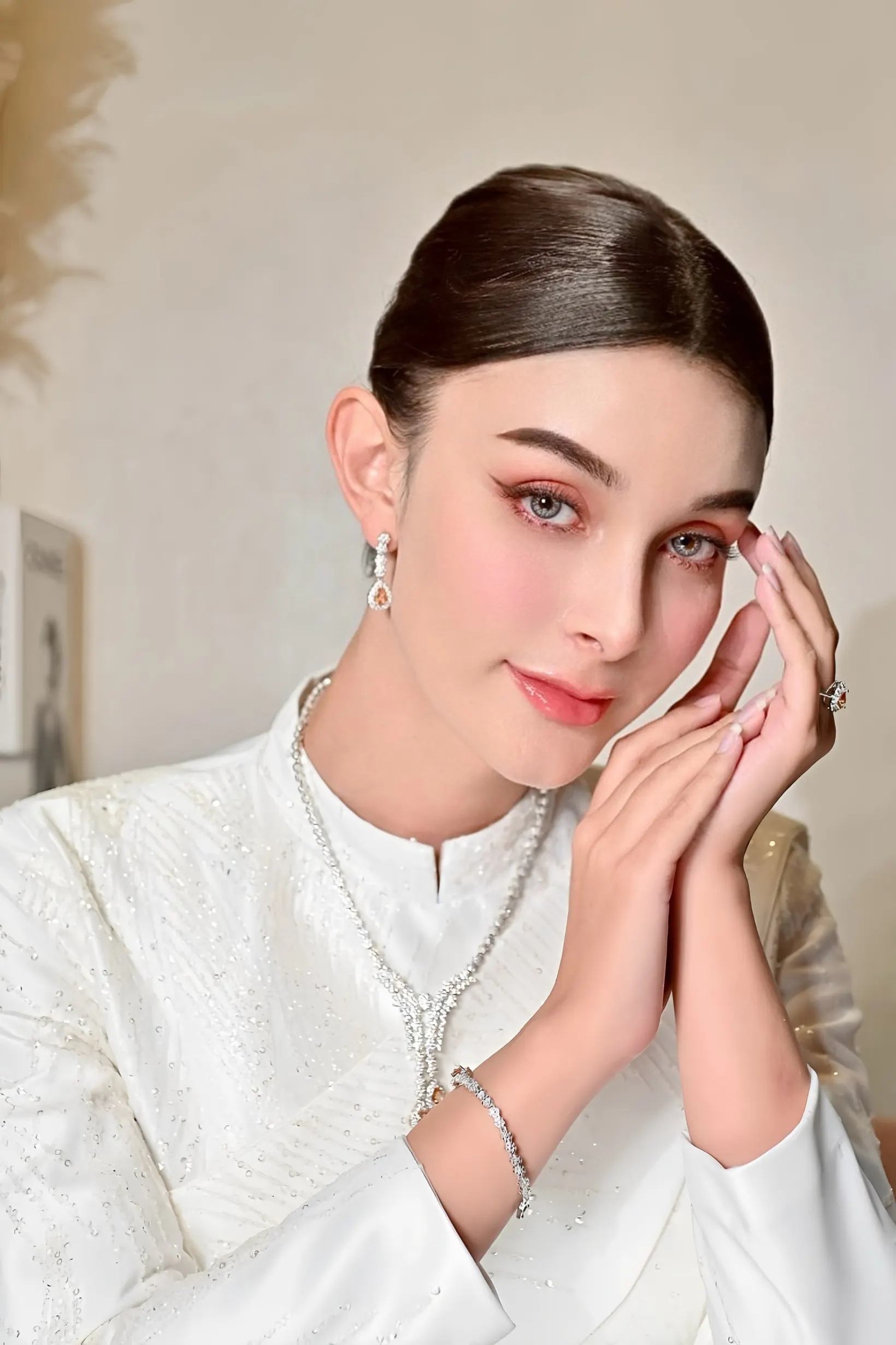 เจ้าสาวในชุดไทยสีขาวปักเลื่อม สวมสร้อยคอเพชร ประสานมือแนบแก้ม แต่งตาโทนส้มพีชและปัดแก้มชมพูอมส้ม ผมรวบเรียบตึง