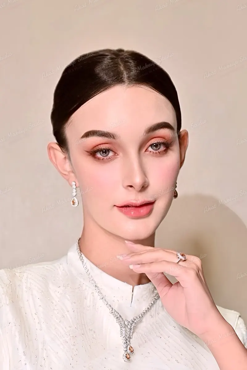 ภาพใกล้ของเจ้าสาวชุดไทยสีขาว ขนตางอนยาว อายแชโดว์โทนพีชทองเข้ากับต่างหูและสร้อยคอ ลิปกลอสชมพูส้ม ผมรวบเรียบ