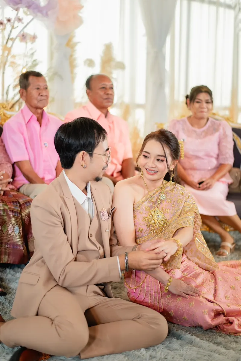 คู่บ่าวสาวนั่งพื้นในพิธีเช้า เจ้าสาวห่มสไบไหมทองสีชมพู แต่งหน้าสดใสยิ้มกว้าง เจ้าบ่าวสวมสูทสีเบจ ญาติผู้ใหญ่นั่งอยู่ด้านหลัง