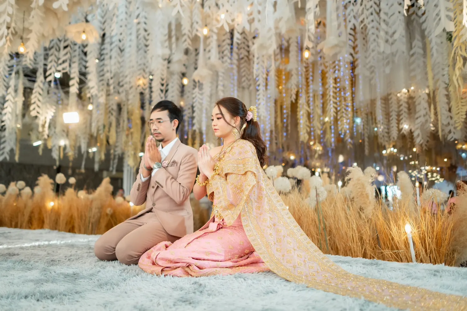 คู่บ่าวสาวนั่งพนมมือบนพรมกลางโถงตกแต่งด้วยใบไม้สีขาวและหญ้าแห้ง เจ้าสาวนุ่งผ้าถุงชมพูห่มสไบทอง ผมเกล้าเก็บเรียบ