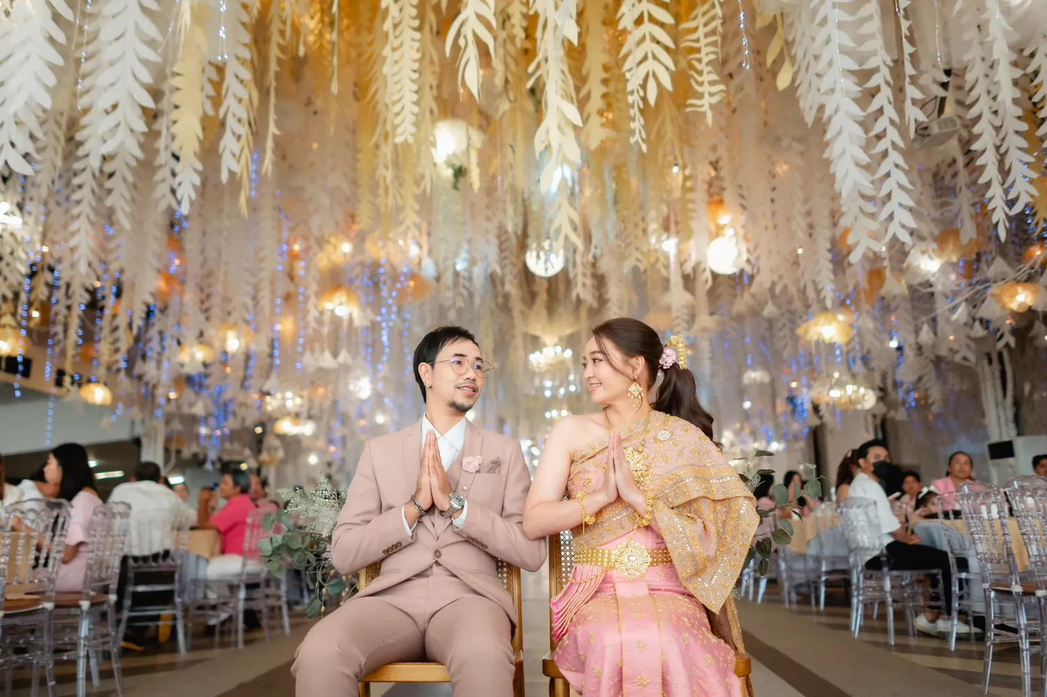 ภาพกว้างคู่บ่าวสาวนั่งพนมมือบนเก้าอี้ในงานแต่งที่ประดับโคมไฟและใบไม้สีขาว เจ้าสาวห่มสไบทองนุ่งผ้าไหมสีชมพู