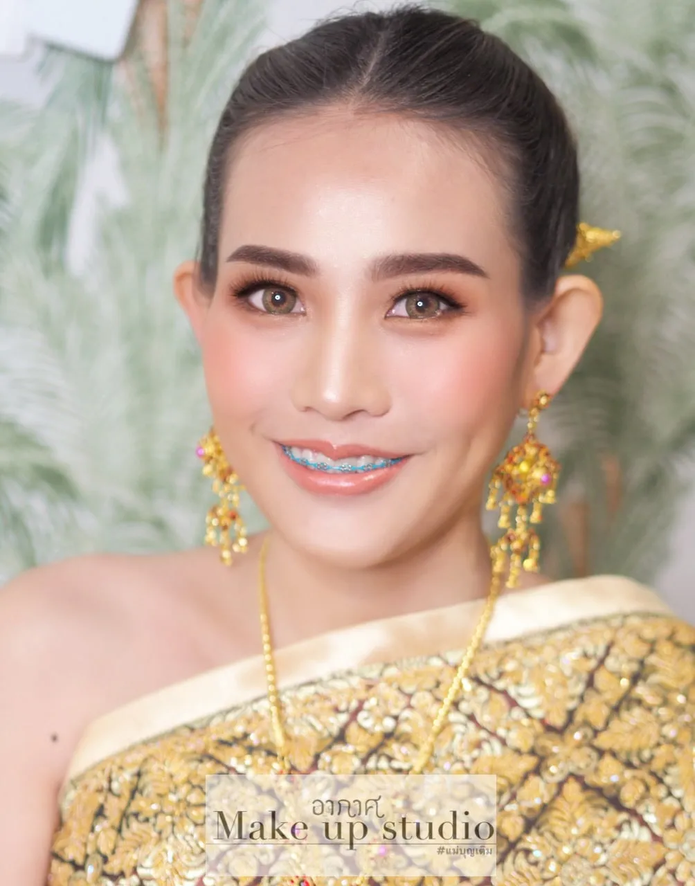 เจ้าสาวชุดไทยสีทองปักลวดลาย ใส่ต่างหูทองห้อยและสร้อยทองเส้นยาว ยิ้มสดใส ผมรวบเรียบ ติดปิ่นทองด้านข้าง มีใบปาล์มสีเขียวอ่อนเป็นฉากหลัง