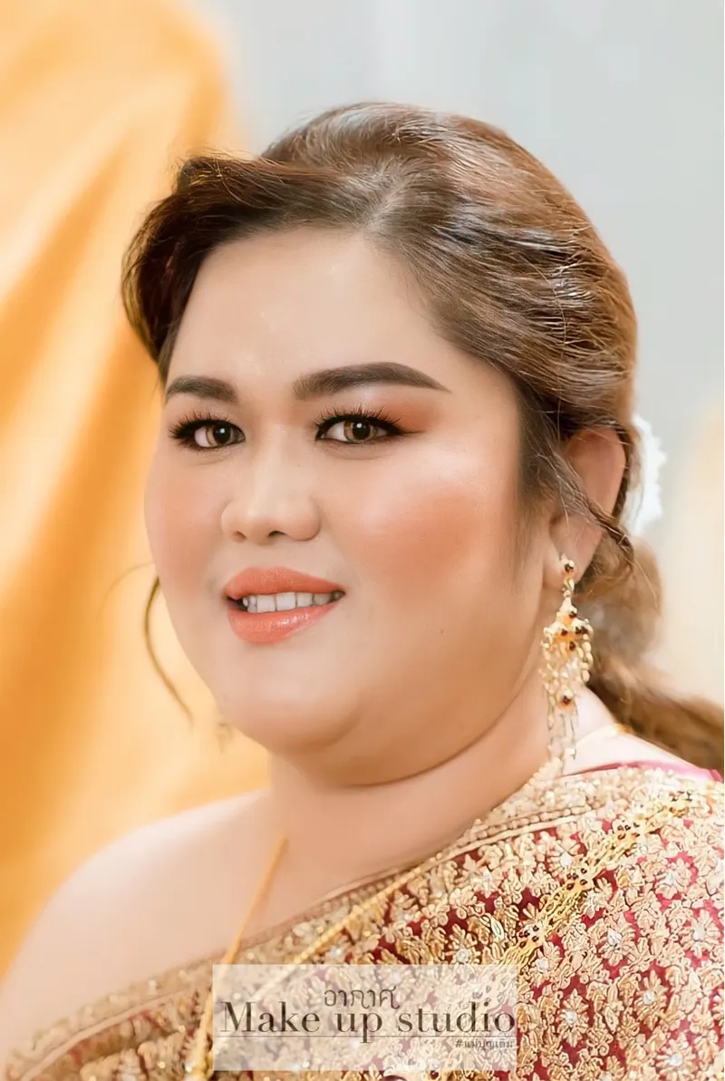 เจ้าสาวชุดไทยสีแดงปักทองกับต่างหูทอง แต่งตาโทนส้มน้ำตาลเน้นขอบตา ปัดแก้มพีช ลิปสีส้มอ่อน เกล้าผมต่ำติดดอกไม้สีขาว