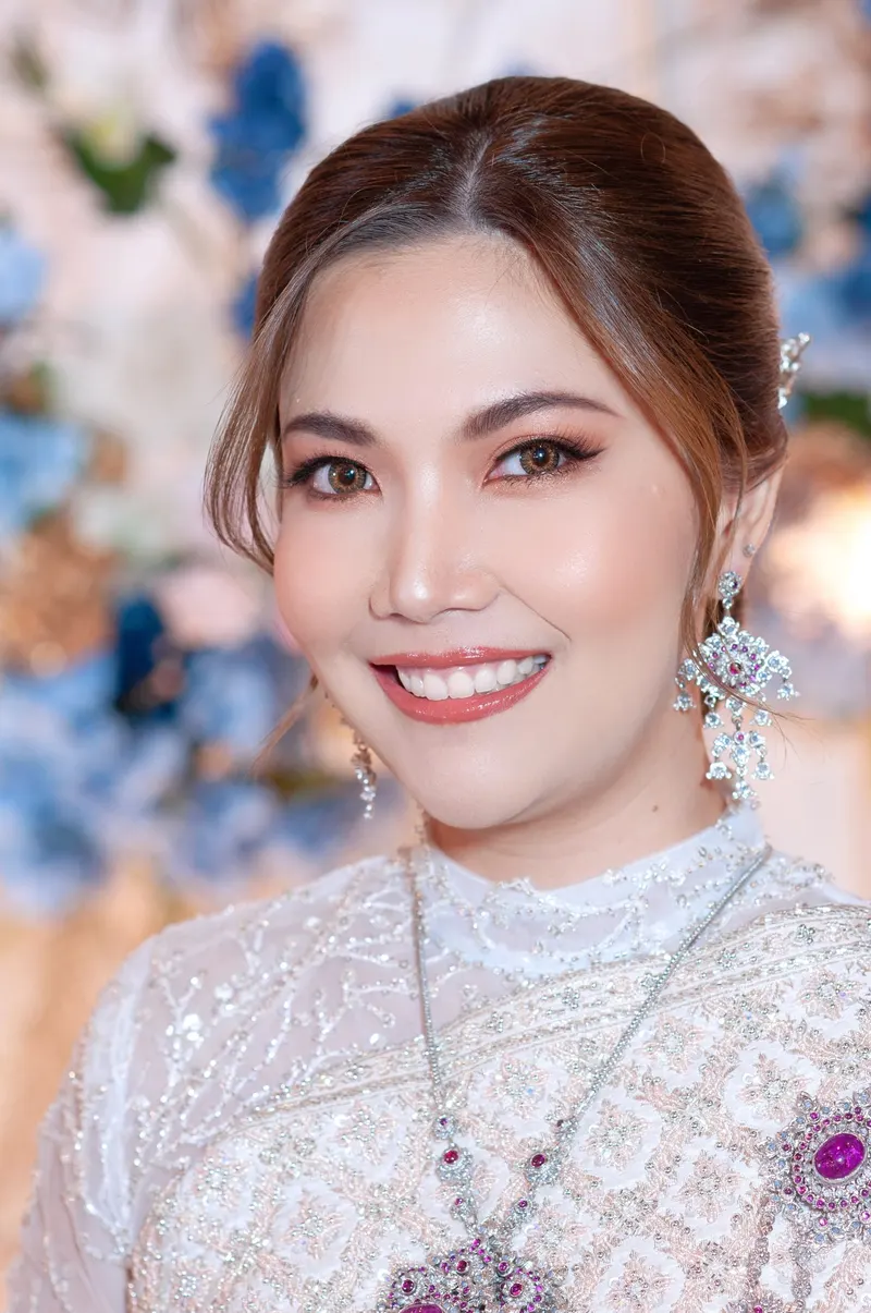 เจ้าสาวชุดไทยลูกไม้สีขาวติดเลื่อมเงิน เครื่องประดับอัญมณีสีม่วง ยิ้มเห็นฟัน แต่งตาโทนส้มทองและลิปสีนู้ดส้ม ผมรวบด้านหลัง