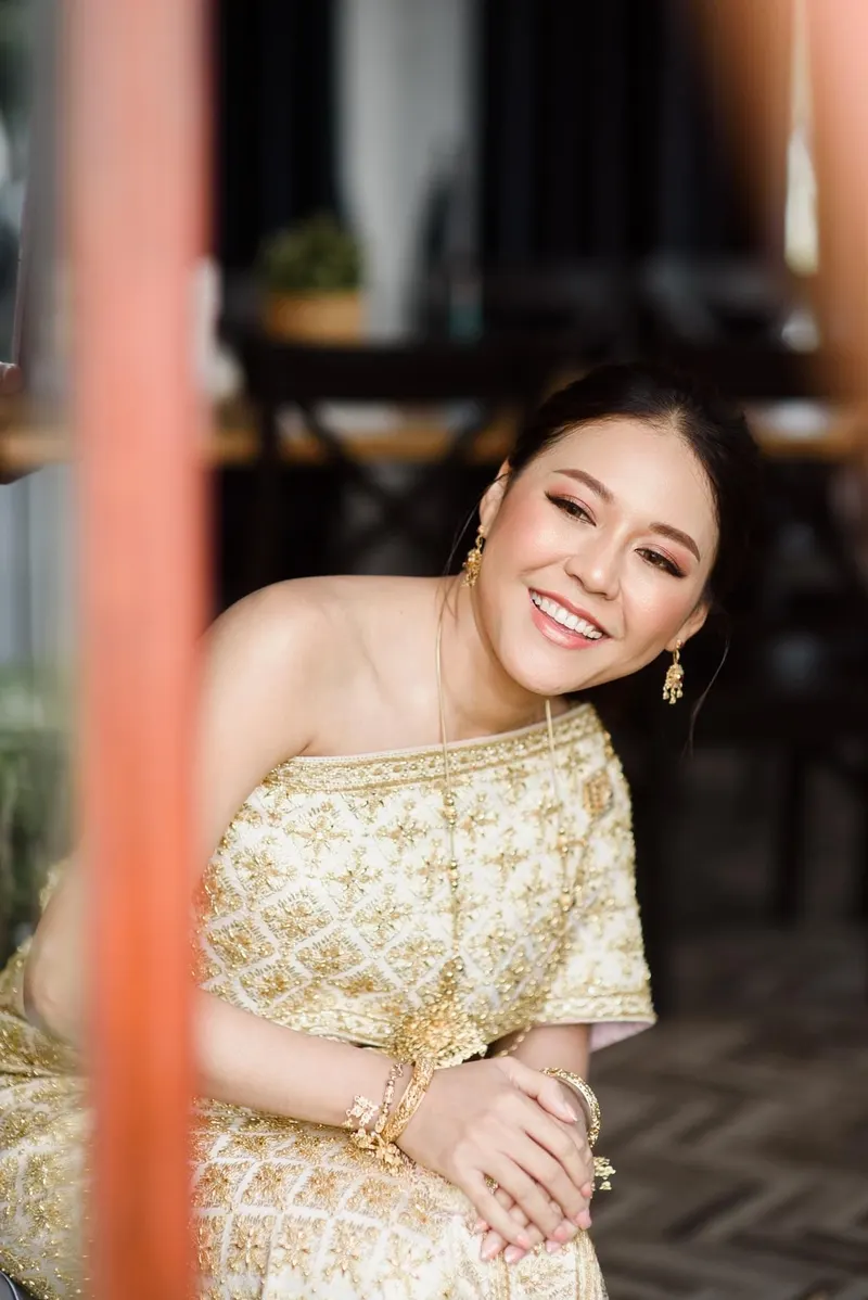 เจ้าสาวนั่งเอียงศีรษะยิ้มกว้างในชุดไทยสีครีมปักลายทอง ใส่ต่างหูทองและกำไลทองหลายวง แต่งตาโทนส้มชมพู ปัดแก้มอมส้ม ผมเกล้าต่ำ มีเสาสีส้มเบลออยู่ด้านหน้า