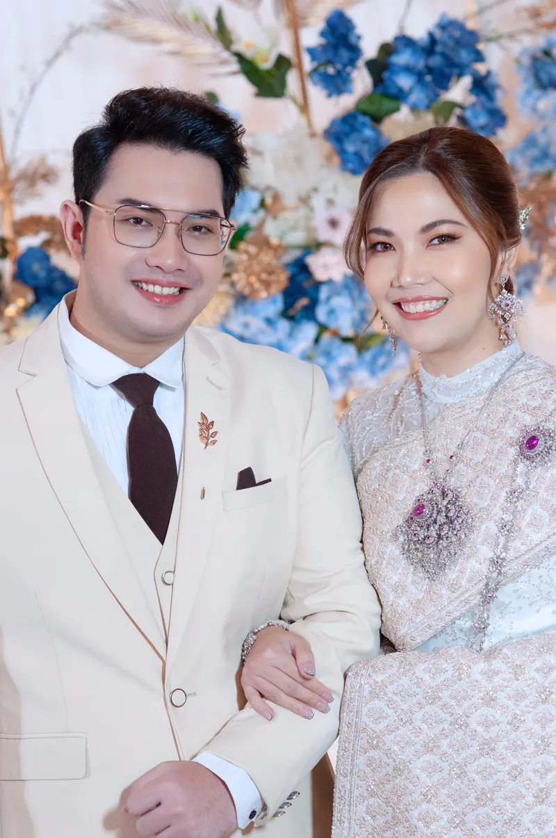 คู่บ่าวสาวยืนเคียงกัน เจ้าสาวชุดไทยลูกไม้ครีมกับเครื่องประดับสีม่วง เจ้าบ่าวสูทครีมใส่แว่น ฉากหลังดอกไม้สีฟ้า แต่งหน้าโทนส้มชมพู