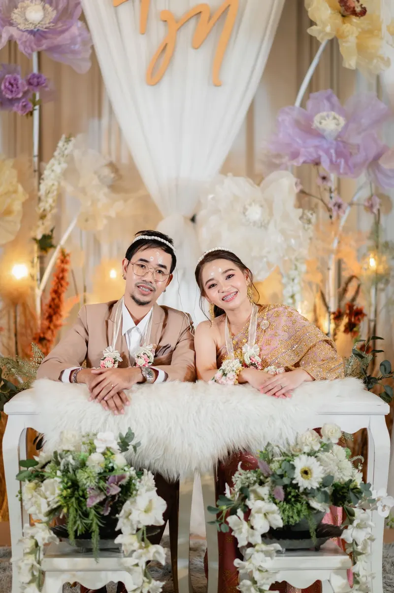 คู่บ่าวสาวนั่งโต๊ะหมั้นบุขนสัตว์สีขาวประดับดอกไม้ เจ้าสาวห่มสไบทองเจ้าบ่าวสูทเบจ ฉากหลังผ้าม่านและดอกไม้กระดาษสีม่วง