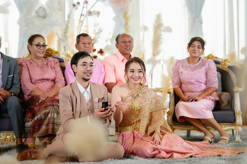 บรรยากาศพิธีเช้า คู่บ่าวสาวนั่งพื้นยิ้มมองไปด้านข้าง ญาติผู้ใหญ่นั่งบนโซฟาด้านหลัง เจ้าสาวห่มสไบทองชมพู