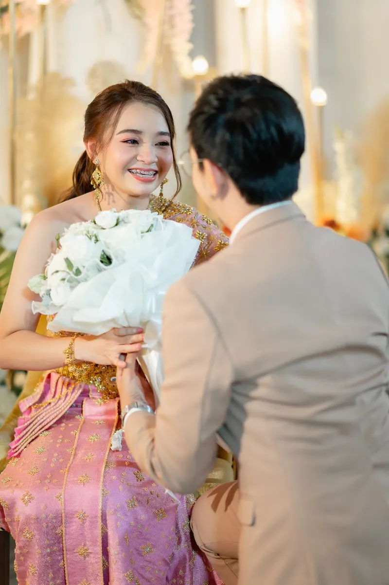 เจ้าสาวชุดไทยสีชมพูถือช่อดอกไม้สีขาว ยิ้มมองเจ้าบ่าวสูทสีเบจที่หันหลังให้กล้อง แสงอบอุ่นฉากหลังประดับดอกไม้