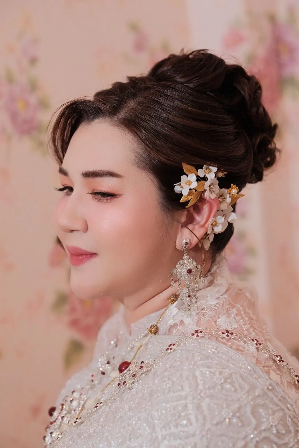 ภาพด้านข้างเจ้าสาวชุดไทยลูกไม้สีขาวปักลูกปัดแดง ผมเกล้าสูงติดดอกไม้ขาวประดับทอง ต่างหูเงินห้อยทับทิม ฉากหลังผนังลายดอกไม้สีชมพู