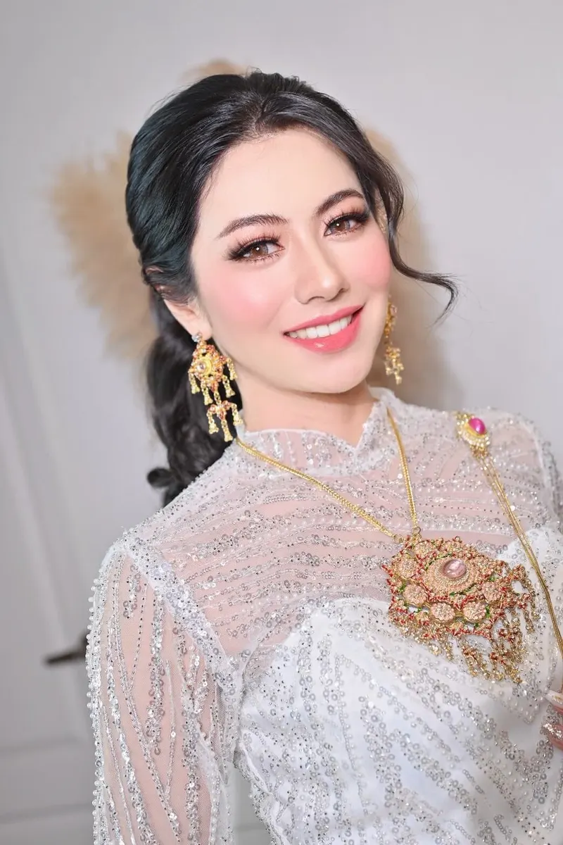 เจ้าสาวชุดไทยลูกไม้ปักเลื่อมสีขาว เข็มกลัดทองอัญมณีสีชมพู ขนตาหนาแต่งตาลายเส้นเฉียง ปัดแก้มชมพูสด ผมรวบข้างลอนปล่อยยาว Akat Makeup Studio